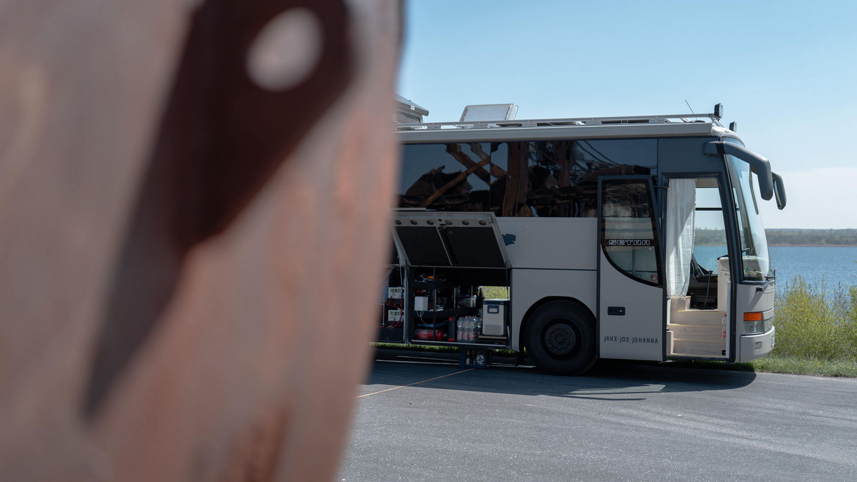 Autocar de tourisme Setra gris transformé en camping-car au bord du lac, porte d’accès et trappes à bagages ouvertes, équipement visible dans l’espace de rangement