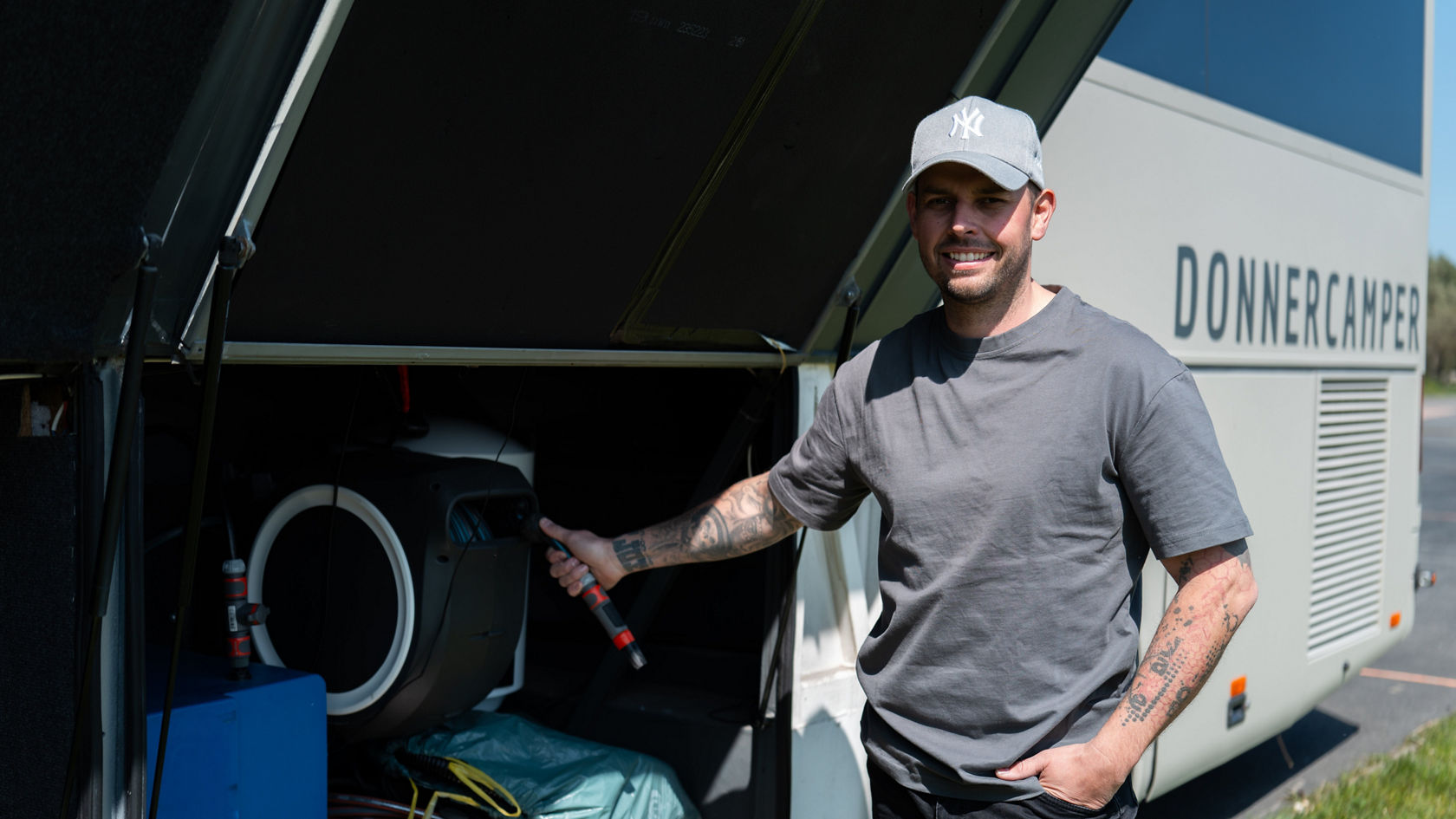 Un homme se tient à côté de l’espace de rangement ouvert d’une transformation de camping-car Setra grise et tient un flexible à côté de l’équipement et des câbles