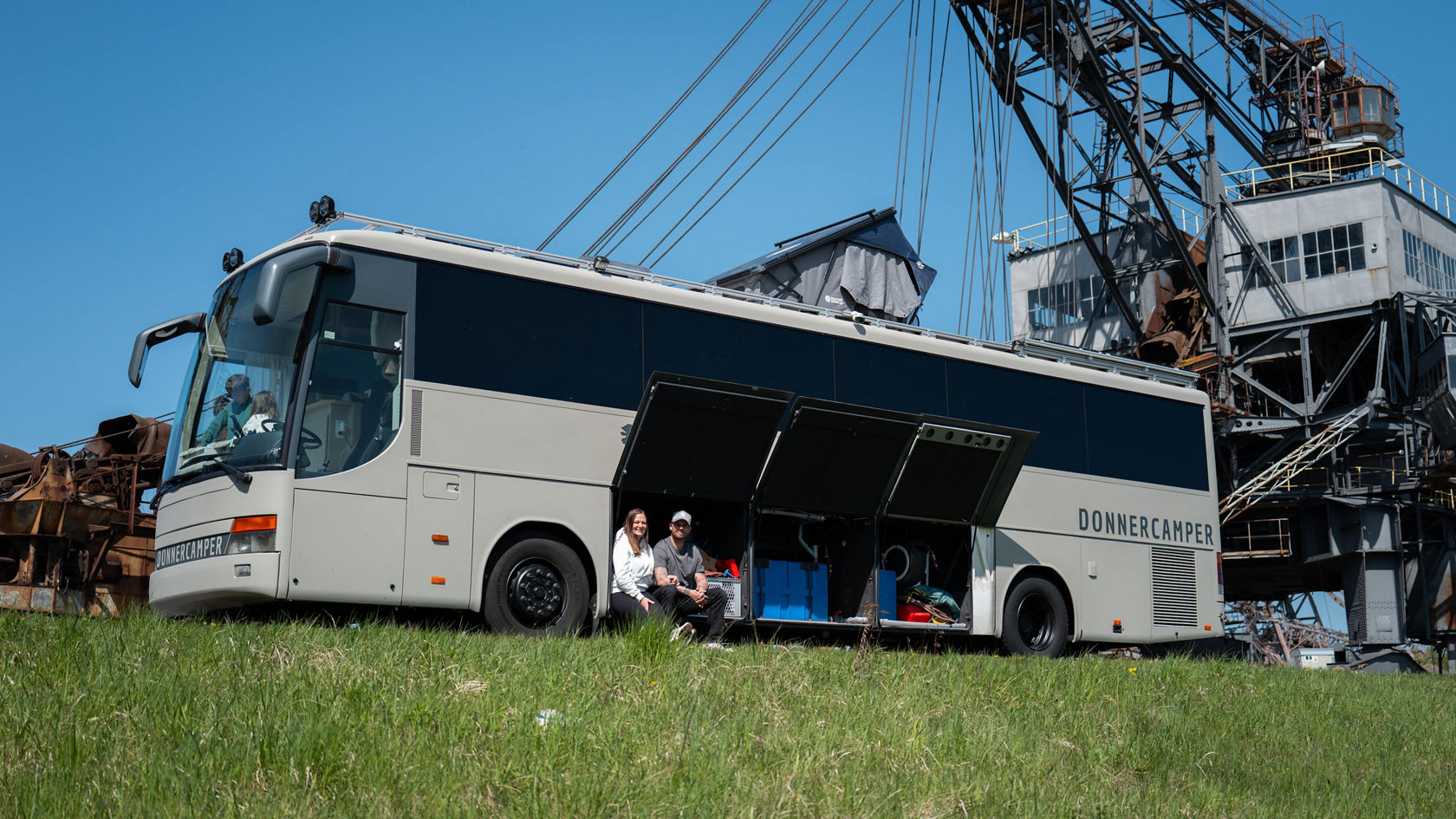 Autocar de tourisme Setra gris transformé en camping-car avec soute à bagages ouverte, deux personnes assises dans la soute à bagages, devant une grue industrielle sous un ciel bleu