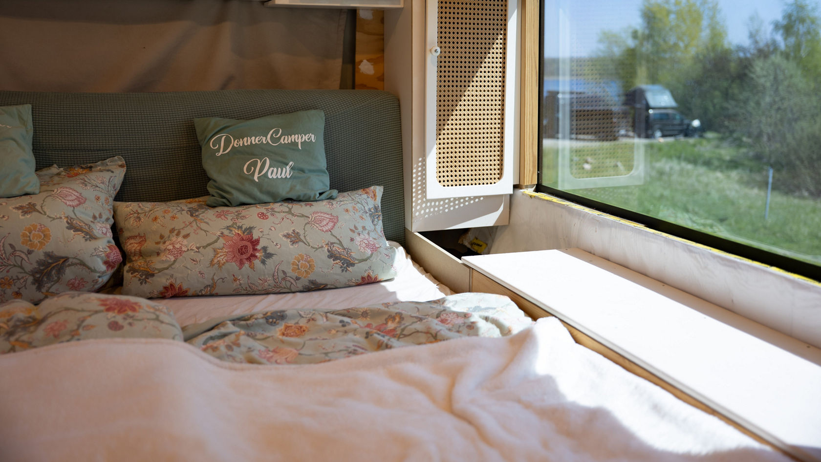 Couchette dans le camping-car Setra transformé : lit double avec linge de lit fleuri, espace de rangement et grande vitre avec vue sur le vert