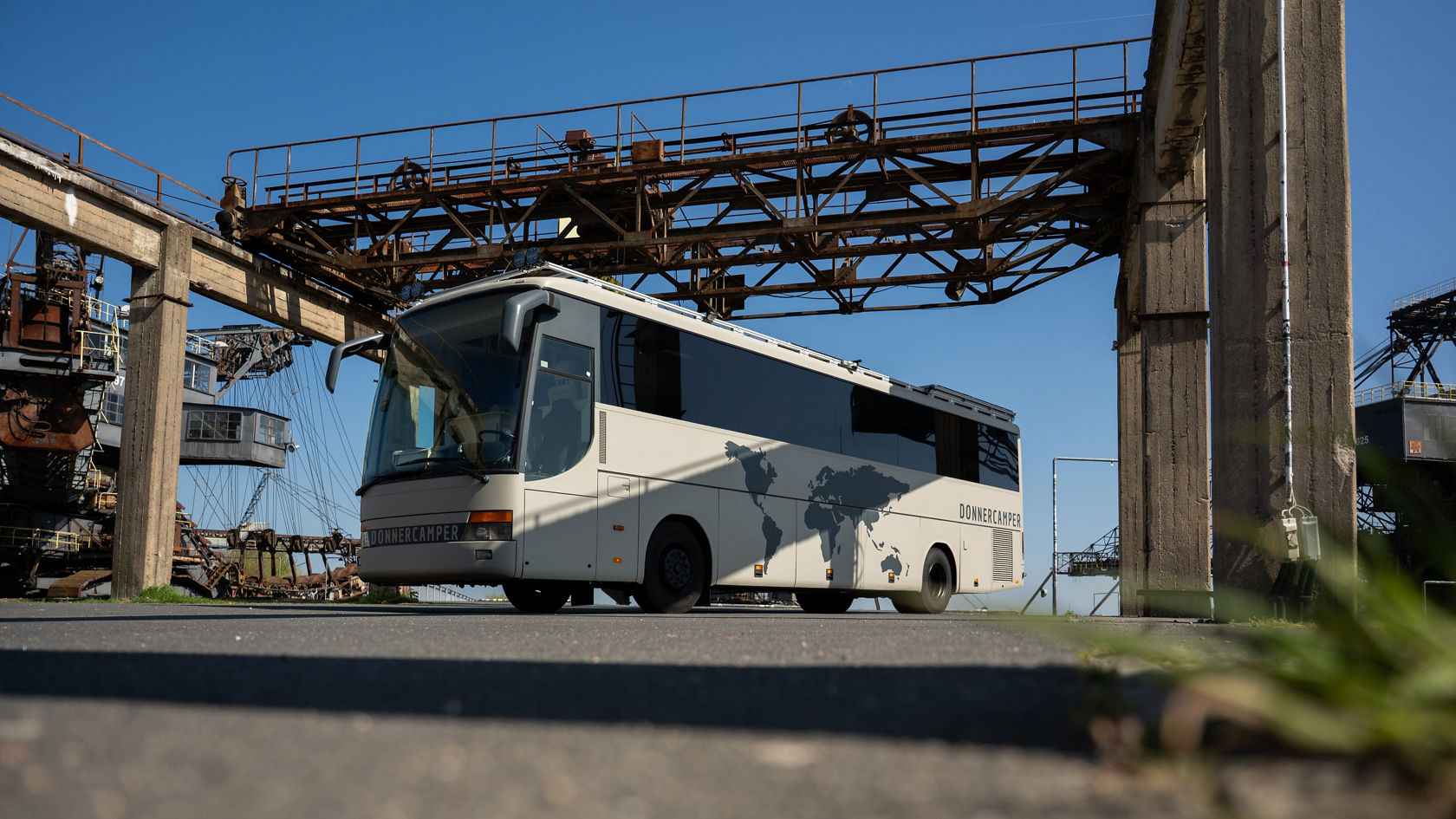 Autocar de tourisme Setra gris transformé en camping-car sous un pont industriel, vue latérale avec cartes du monde sur la superstructure