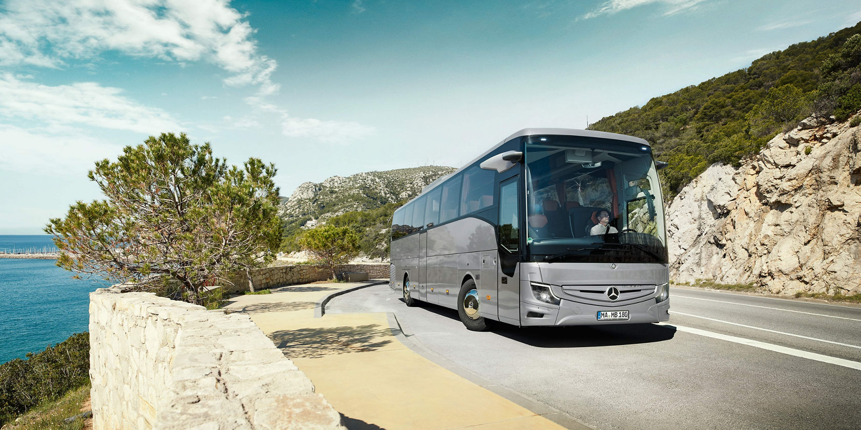 Mercedes‑Benz Tourismo Reisebus fährt auf kurviger Küstenstraße entlang felsiger Hänge, Meerblick unter blauem Himmel mit ein paar Wolken