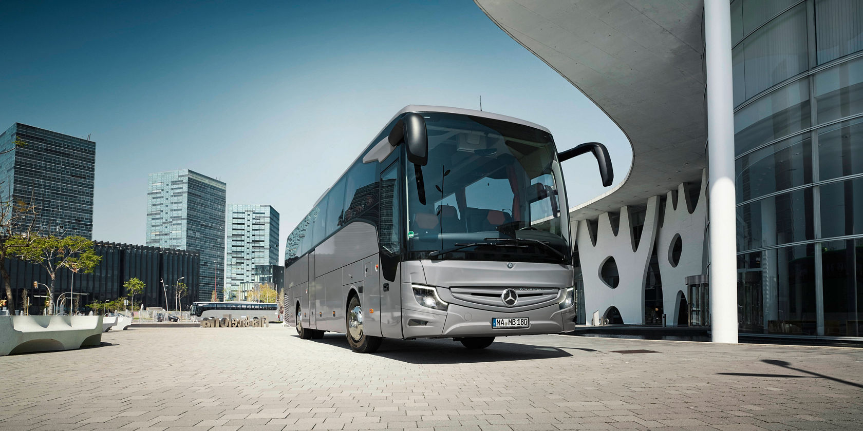 Silberner Mercedes‑Benz Tourismo Reisebus vor moderner Glasfassade, Frontansicht auf gepflasterter Fläche in der Stadt