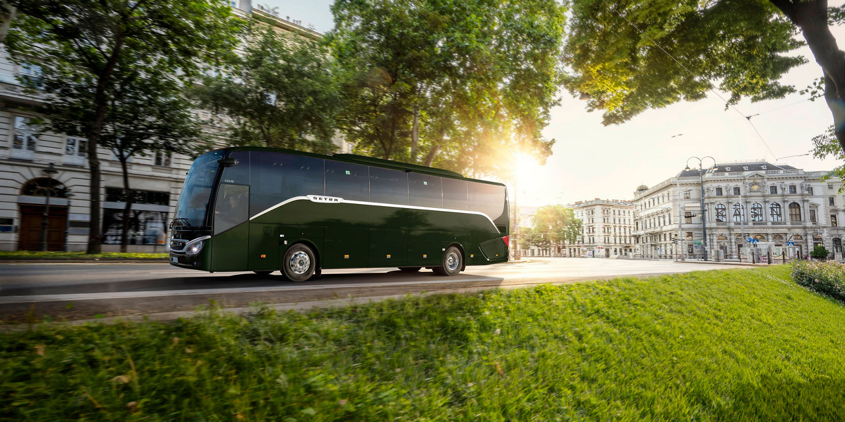 Autocarro de turismo Setra ComfortClass verde a percorrer uma avenida urbana ao pôr do sol, com árvores e edifícios históricos em segundo plano.