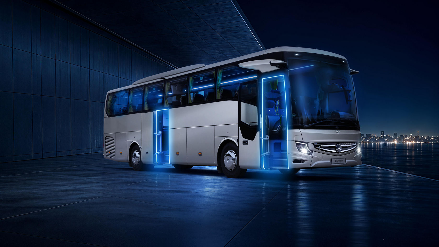 Mercedes‑Benz Tourismo Reisebus bei Nacht, mit blau beleuchteten Einstiegen und geöffneten Türen, im weiter Ferne Lichtsilhouette einer Stadt