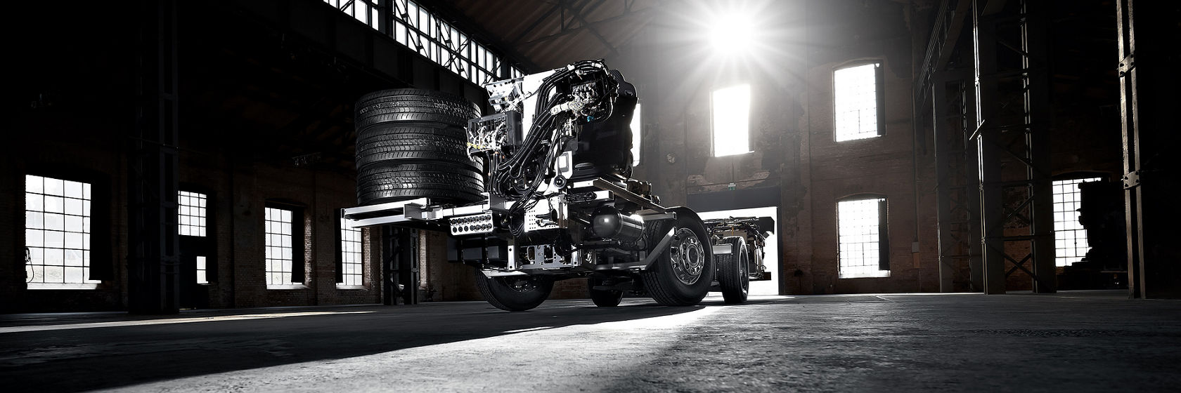 Mercedes‑Benz Bus‑Chassis mit freiliegendem Heckmodul und gestapelten Reifen in Industriehalle, Gegenlichtaufnahme